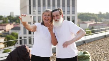 Yaşlı çift aydınlık güneşli bir günde çatıda birlikte selfie çekerek neşe ve dostluk gösteriyorlar.