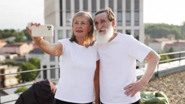 Yaşlı bir çift çatıda durmuş kucaklaşıyorlar, selfie çekiyorlar ve gülümsüyorlar. Şehir geçmişi yeşillik, günlük atmosfer, beyaz gömlekler. Kavramlara aşk, birliktelik de dahil.