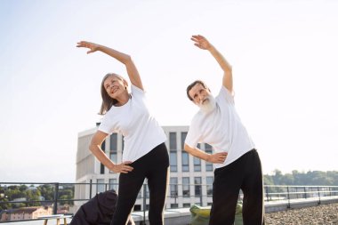 Yaşlı adam ve kadın, açık havada, çatıda, parlak güneş ışığında esneme egzersizleri yapıyorlar. Sağlık, hareketlilik, günlük aktivite, mutlu yaşam tarzı vurgulanıyor.