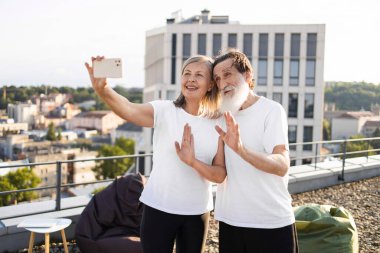 Sakallı yaşlı bir adam ve beyaz tişörtlü bir kadın çatıda selfie çekiyorlar. Şehir çevresi arkada görünür. Mutlu ve rahat görünüyorlar, anılarını yakalarken dışarıda eğleniyorlar..