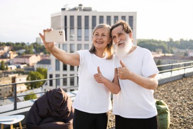 Yaşlı adam ve kadın dışarıda selfie çekerken gülümsüyor, başparmaklarını kaldırıyor, altın çağlarda aktif yaşam tarzını ve pozitifliği destekliyor..
