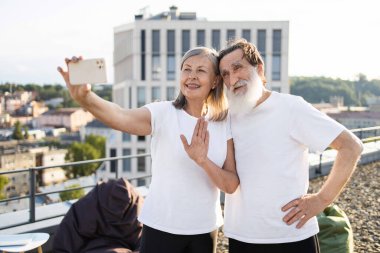 Yaşlı çift aydınlık güneşli bir günde çatıda birlikte selfie çekerek neşe ve dostluk gösteriyorlar.