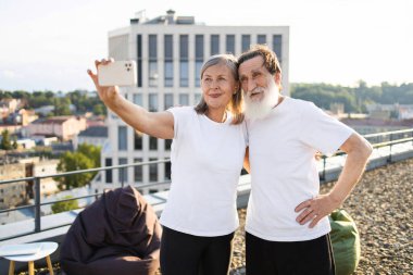 Yaşlı bir çift çatıda durmuş kucaklaşıyorlar, selfie çekiyorlar ve gülümsüyorlar. Şehir geçmişi yeşillik, günlük atmosfer, beyaz gömlekler. Kavramlara aşk, birliktelik de dahil.