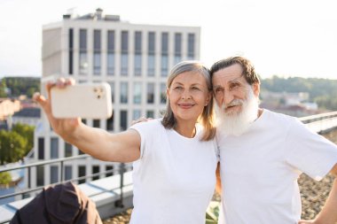 Sakallı yaşlı bir adam ve güneş ışığında çatıda selfie çeken yaşlı bir kadın. Şehir ortamında mutluluk ve birliktelikle yakalanan sıradan bir açık hava anı.