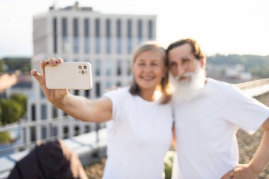 Sakallı yaşlı bir adam ve güneşli bir günde şehir ortamında selfie çeken yaşlı bir kadın, günlük beyaz gömlekler giyiyor, neşe ve beraberliği ifade ediyor, dışarıda eğleniyor, çatı arkaplanından keyif alıyor.