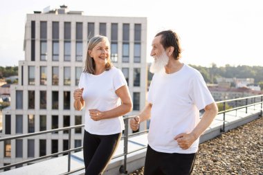 Aktif kıdemli erkek ve kadın çatıda koşuyor, sağlıklı yaşam tarzı ve uygunluğu gösteriyor.