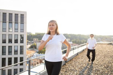 Yaşlı kadın çatıda koşu yaparak aktif kalıyor, yanında şehir manzarasında sakallı bir adam var. Sağlığı, zindeliği, canlılığı teşvik eder..
