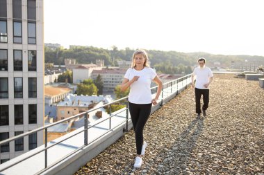 Üst düzey erkek ve kadın birlikte çatıda egzersiz yapıyorlar. Etrafı güneş ışığıyla çevrili.