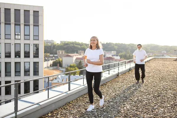 Üst düzey erkek ve kadın çatıda koşuyor, spor salonunun ve şehir ortamının tadını çıkarıyorlar. Spor kıyafetleri giyiyorlar ve mutlu ve motive görünüyorlar. Sağlıklı ve aktif bir yaşam tarzlarını yansıtıyorlar..