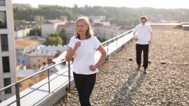 Üst düzey erkek ve kadın çatıda koşuyor, spor salonunun ve şehir ortamının tadını çıkarıyorlar. Spor kıyafetleri giyiyorlar ve mutlu ve motive görünüyorlar. Sağlıklı ve aktif bir yaşam tarzlarını yansıtıyorlar..