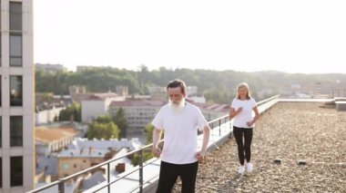 Beyaz gömlekli son sınıf öğrencisi şehrin çatısında egzersiz yapıyor. Arka plandaki kadın da fitness aktivitelerine katılıyor. Manzaralı şehir manzarası dinamik ve canlandırıcı bir atmosfer yaratır.