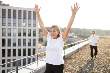 Aktif giyimli yaşlı kadın şehir çatısı üzerinde kollarını kaldırarak, dışarıda neşe ve canlılık ifade ediyor..