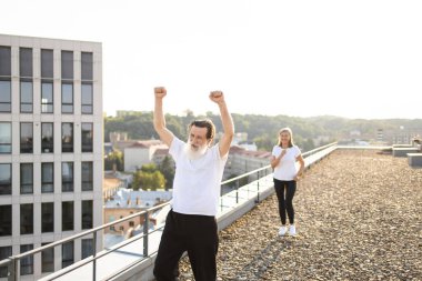 Beyaz gömlekli son sınıf öğrencisi şehrin çatısında egzersiz yapıyor. Arka plandaki kadın da fitness aktivitelerine katılıyor. Manzaralı şehir manzarası dinamik ve canlandırıcı bir atmosfer yaratır.