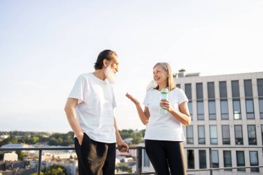 Spor kıyafetleri içinde yaşlı bir adam ve kadın çatıda dikiliyor, spor yaptıktan sonra neşe ve rahatlama ifade ediyorlar..