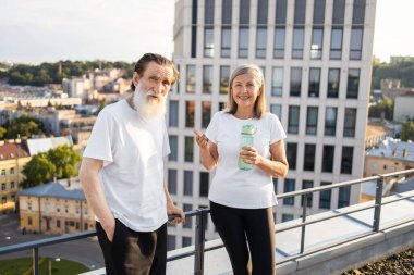 İki yaşlı yetişkin, erkek ve dişi, güneşli şehir merkezinde sabah egzersizlerinden sonra çatıda duruyorlar..