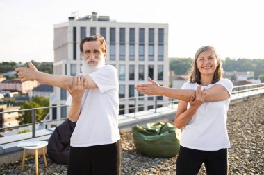 Yoga sırasında dışarıda kol germe egzersizi yapmaktan hoşlanan aktif kıdemli erkek ve kadın. İkisi de günlük giysi giyiyor. Arkaplanda şehrin ufuk çizgisi ve kentsel ortam, sağlık bilincinin aktarılması yer alıyor