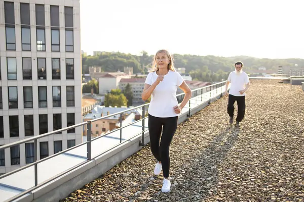Yaşlı bir adam ve kadın gün doğumunda çatıda koşuyor. Sağlık ve zindeliği destekliyor. Aktif kıdemli çift, takım çalışmasını ve sağlıklı yaşam tarzını vurgulayan günlük kıyafetlerle dışarıda egzersiz yapmaktan hoşlanıyor.