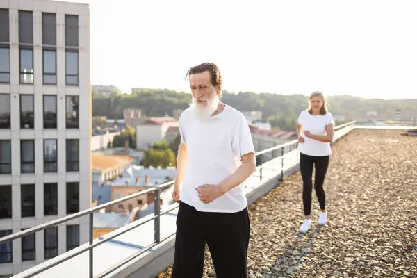 Üst düzey erkek ve kadın, şehrin çatısında koşuyor. Kentsel yaşlılar arasında spor ve refahı destekleyen aktif yaşam tarzı, sağlık ve refah. Güneş ışığı şehir manzarasını güçlendirir ve sahneye sıcaklık katar.