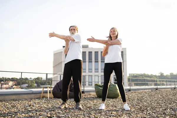 Üst düzey erkek ve kadın çatıda esneme egzersizleri yapıyor. Görüntü, denge esnekliği ve refahı vurgulayan yaşlı yetişkinler arasında fitness sağlıklı alışkanlıkları ve aktif yaşam tarzını teşvik eder.