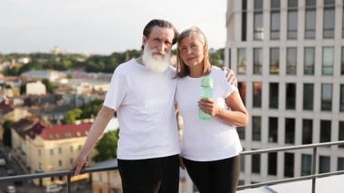 Yaşlı adam ve kadın sabah egzersizinden sonra çatıda durup gülümsüyor, enerji, sağlık ve arkadaşlık gösteriyorlar. Zindelik ve refah kavramına sahip modern kentsel ortam.