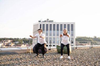 Yaşlı adam ve kadın sıcak ve huzurlu bir sabahta çatıda çömelme hareketleri yaparak yaşlılara aktif yaşam tarzını tanıtıyorlar..