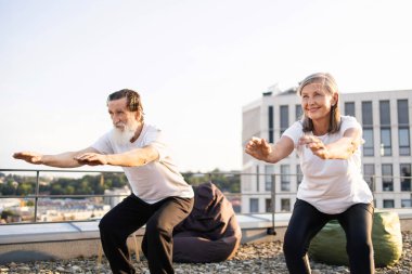Yaşlı adam ve kadın çatı terasında birlikte egzersiz yapıyor, çömelme ve fiziksel sağlığa odaklanıyor. Gelişmiş fitness ve refah için aktif yaşam tarzına girmek.