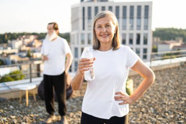 Üst düzey bir kadın çatıda elinde su şişesi, gülümseme, spor anlayışı, sağlık. Arka planda açık çatı ve adam görünüyor. Olgun ve yaşlı yetişkinler için aktif açık hava yaşam tarzını teşvik eder.