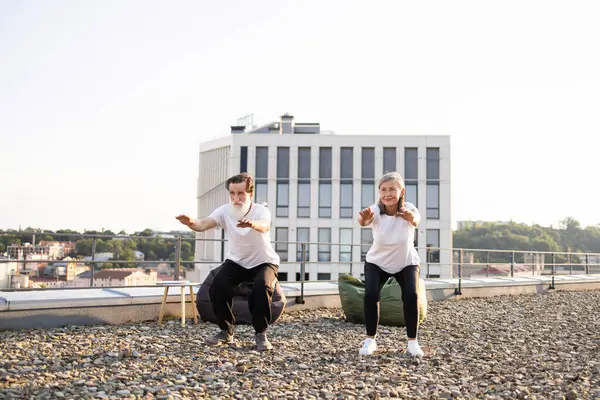 Yaşlı adam ve kadın sıcak ve huzurlu bir sabahta çatıda çömelme hareketleri yaparak yaşlılara aktif yaşam tarzını tanıtıyorlar..