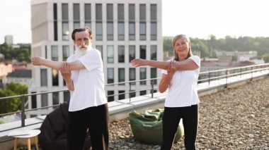 Yoga sırasında dışarıda kol germe egzersizi yapmaktan hoşlanan aktif kıdemli erkek ve kadın. İkisi de günlük giysi giyiyor. Arkaplanda şehrin ufuk çizgisi ve kentsel ortam, sağlık bilincinin aktarılması yer alıyor