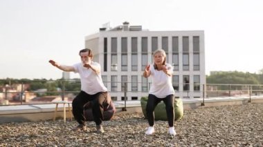 Yaşlı adam ve kadın sıcak ve huzurlu bir sabahta çatıda çömelme hareketleri yaparak yaşlılara aktif yaşam tarzını tanıtıyorlar..