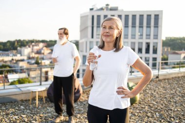 Kıdemli adam ve kadın dışarıda mola veriyorlar, su şişeleriyle su içiyorlar, şehir çatısı arka planı var. Manzaralı şehir ortamında sağlıklı, aktif yaşam tarzının ve açık hava ortamının tadını çıkarıyorum..