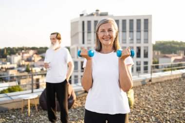 Şehrin çatısında mavi dambıllar tutan gülümseyen son sınıf öğrencisi bir kadın, şehir dışı aktivitesi. Formunu, sağlığını, sağlığını temsil ediyor. Arka plandaki yaşlı adam açık havada eğleniyor.