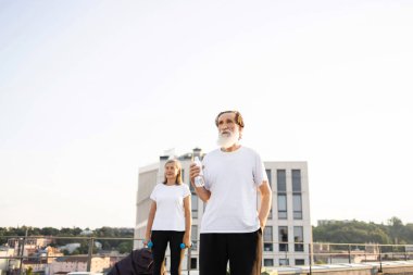 Yaşlı adam ve kadın halterler ve su şişeleriyle dışarıda egzersiz yapıyor, aktif yaşam tarzının tadını çıkarıyorlar. Güneşli bir günde açık şehir ortamında, yetişkinlikte sağlık, zindelik ve canlılık vurgulanıyor.