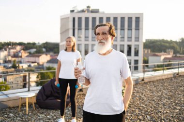Beyaz sakallı, elinde su şişesi olan yaşlı bir adam, çatıda beyaz gömlek giyen bir kadın. Manzaralı kentsel arka plan, sağlıklı fitness rutinine odaklan, molada su hidrasyonu.