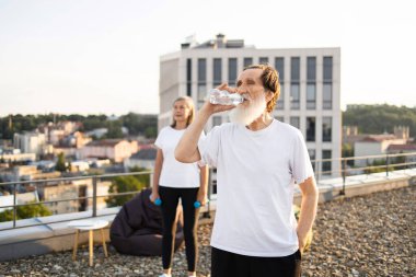 Ağırlık tutan kadın dışarıda beklerken beyaz sakallı, su içen yaşlı bir adam. Çatıda spor yaparken çekilen sahne su ve egzersizin önemini gözler önüne seriyor..