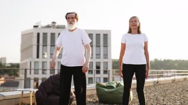 Yaşlı adam ve kadın çatı terasında birlikte egzersiz yapıyor, çömelme ve fiziksel sağlığa odaklanıyor. Gelişmiş fitness ve refah için aktif yaşam tarzına girmek.