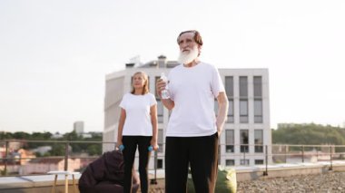 Ağırlık tutan kadın dışarıda beklerken beyaz sakallı, su içen yaşlı bir adam. Çatıda spor yaparken çekilen sahne su ve egzersizin önemini gözler önüne seriyor..