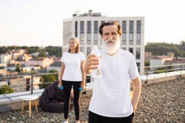 Beyaz sakallı yaşlı bir adam çatıda su şişesi tutuyor ve yaşlı bir kadın egzersiz yapıyor. Açık hava fitness, hidrasyon, refah. Yaşlı yetişkinler için parlak kentsel yaşam biçimi.