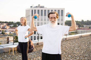 Yaşlı bir adam ve kadın dambıllarla çatıda birlikte egzersiz yapıyorlar. Sağlıklı yaşam tarzı etkinliği, açık hava ortamındaki yaşlı yetişkinler için spor, denge ve refahı vurguluyor..