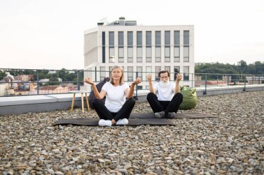 İki yaşlı birey, kadın ve erkek, Lotus 'ta poz veriyorlar. Yoga sırasında çatıdaki kentsel alanda poz veriyorlar. Sağlık, rahatlama ve zindelik vurguluyorlar.