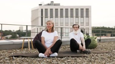 İki yaşlı birey, kadın ve erkek, Lotus 'ta poz veriyorlar. Yoga sırasında çatıdaki kentsel alanda poz veriyorlar. Sağlık, rahatlama ve zindelik vurguluyorlar.