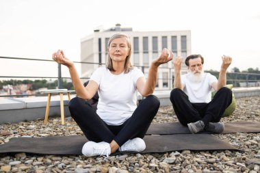 Yaşlı beyaz çift çatıda bacak bacak üstüne atarak oturuyor, meditasyon yapıyor ve yoga yapıyorlar, rahatlama gösteriyorlar..