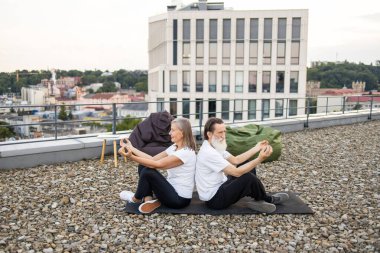 Üst düzey erkek ve kadın nilüfer çiçeğiyle yoga yapıyor çatıda poz veriyor rahatlama ve zindeliği birleştiriyor. Arka planda şehir manzarası görünüyor. Mankenler, sağlıklı yaşlanmayı temsil eden yaşlı erkek ve dişilerdir.