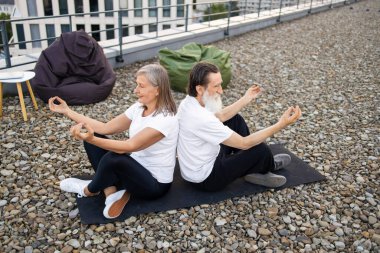Kıdemli çift, arka arkaya dışarıda Lotus pozu vererek rahatlatıcı meditasyon yapıyor. Kadın ve erkek, siyah ve beyaz giysi giyiyor. Kentsel çevrede dengeli yaşam tarzı ve uyum kavramı.