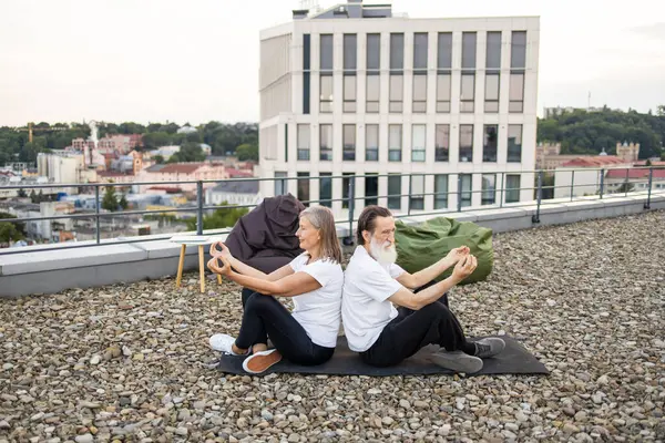 Üst düzey erkek ve kadın nilüfer çiçeğiyle yoga yapıyor çatıda poz veriyor rahatlama ve zindeliği birleştiriyor. Arka planda şehir manzarası görünüyor. Mankenler, sağlıklı yaşlanmayı temsil eden yaşlı erkek ve dişilerdir.