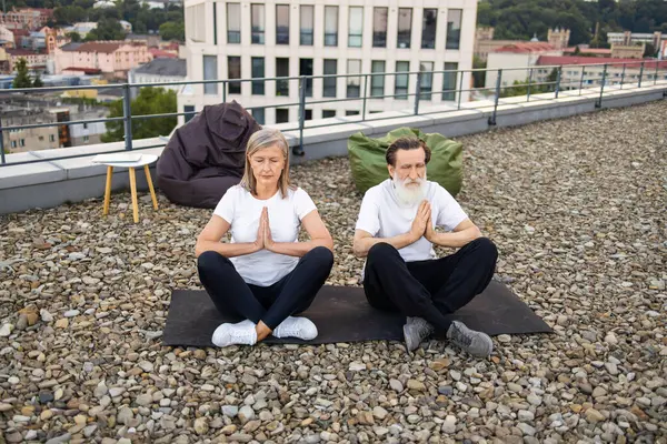 Üst düzey kadın ve erkek yoga yapıyor. Çatıdaki yoga minderlerinde poz veriyorlar..