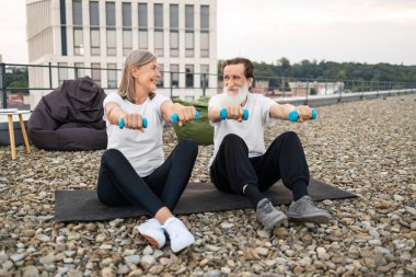 Yaşlı bir adam ve kadın çatıdaki minderlerde oturmuş halterlerle spor yapıyorlar..