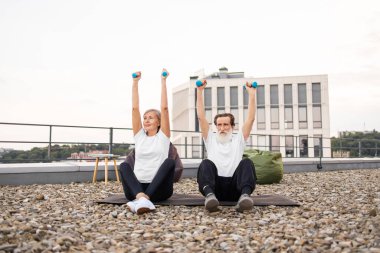 Yaşlı bir adam ve kadın spor yaparken dambılları kaldırıyor. Terasta oturuyorlar. Arka planda modern bir bina var.