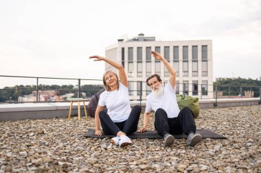 Yaşlı erkek ve kadın yoga yaparken çatıda birlikte geriliyor, gülümsüyor ve açık hava spor faaliyetlerinden keyif alıyor, sağlıklı yaşam tarzını ve aktif yaşlanmayı vurguluyor..