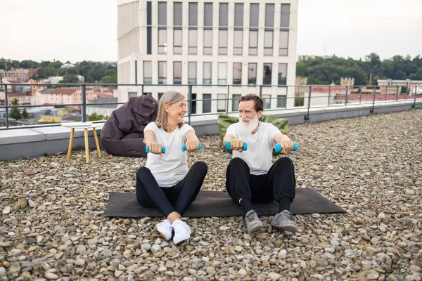 Üst düzey çift dambıllarıyla çatıda egzersiz yapıyor. Yaşlı adam ve kadın dışarıda spor yapıyorlar..